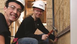 Two students wearing hard hats and holding a hammer smile at the camera