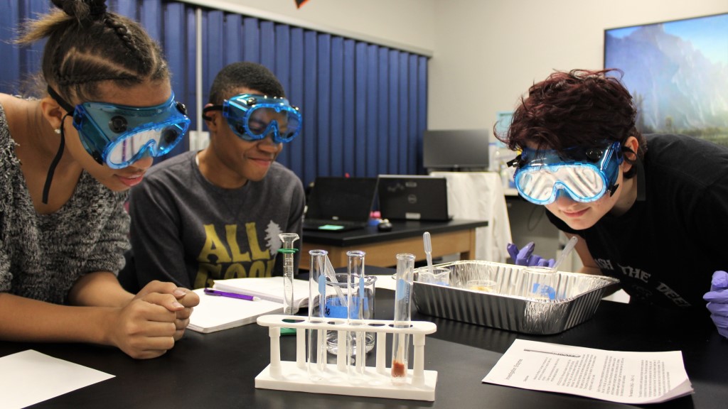 Three students look at test tubes while wearing safety glasses