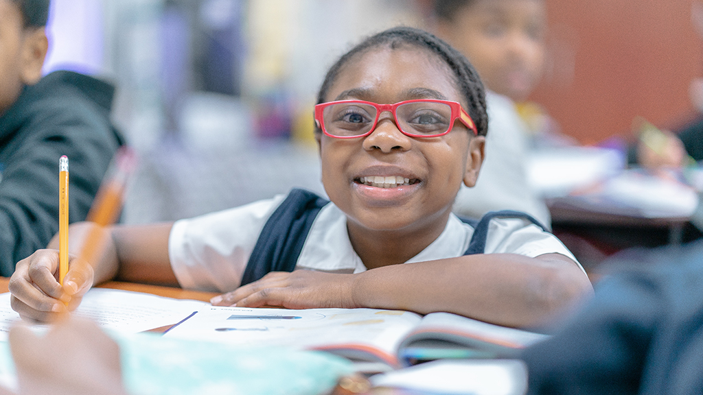 a student with big red glasses smiles brightly at the camera as she works at her desk