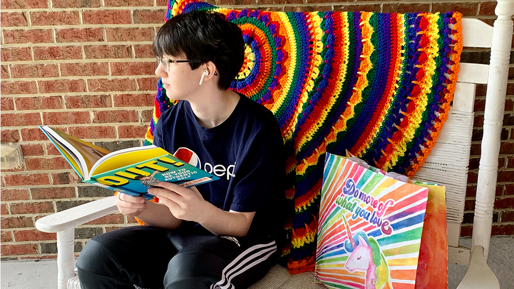 a student sits on a chair with a rainbow blanket and reads a book entitled "out!"