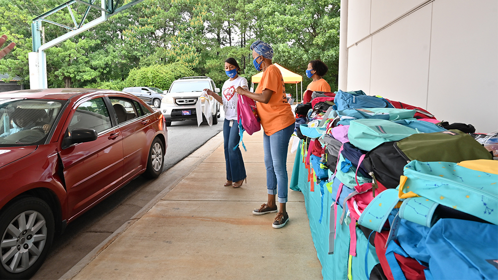 a large row of backpacks are being handed out by volunteers to cars in a drive thru