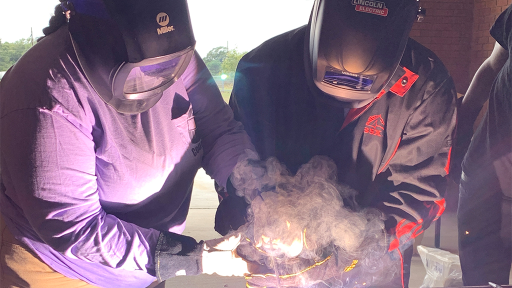two students wearing protective clothes use wielding equipment, producing a cloud of steam and light 