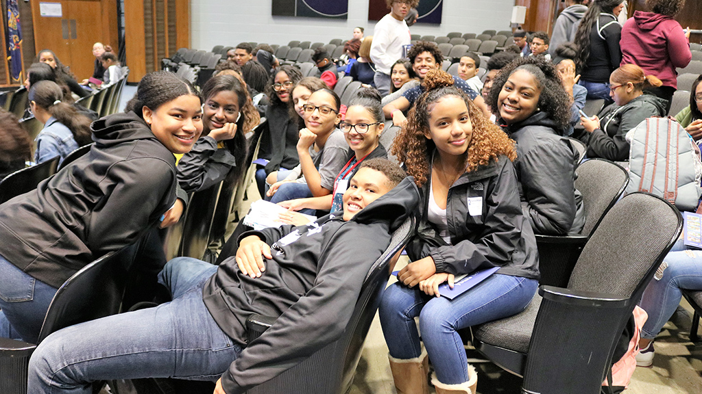a group of students sit in an auditorium, they are smiling and laughing at the camera