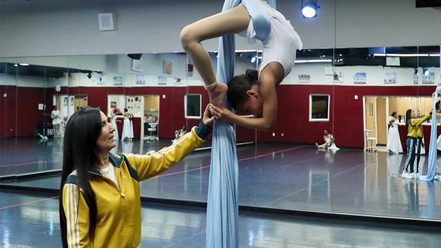 an instructor and a dancer, who hangs suspended from a sheet attached to the ceiling 