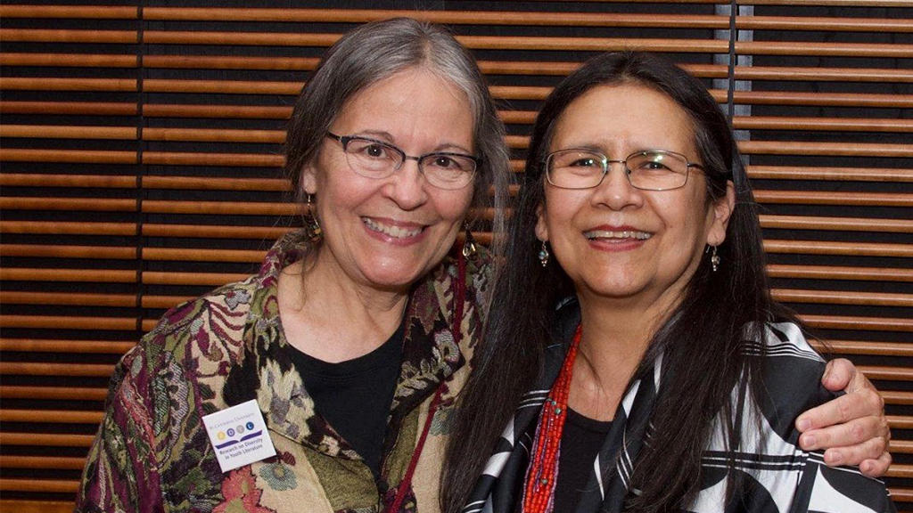 Authors Jean Mendoza and  Debbie Reese stand shoulder-to-shoulder and smile at the camera