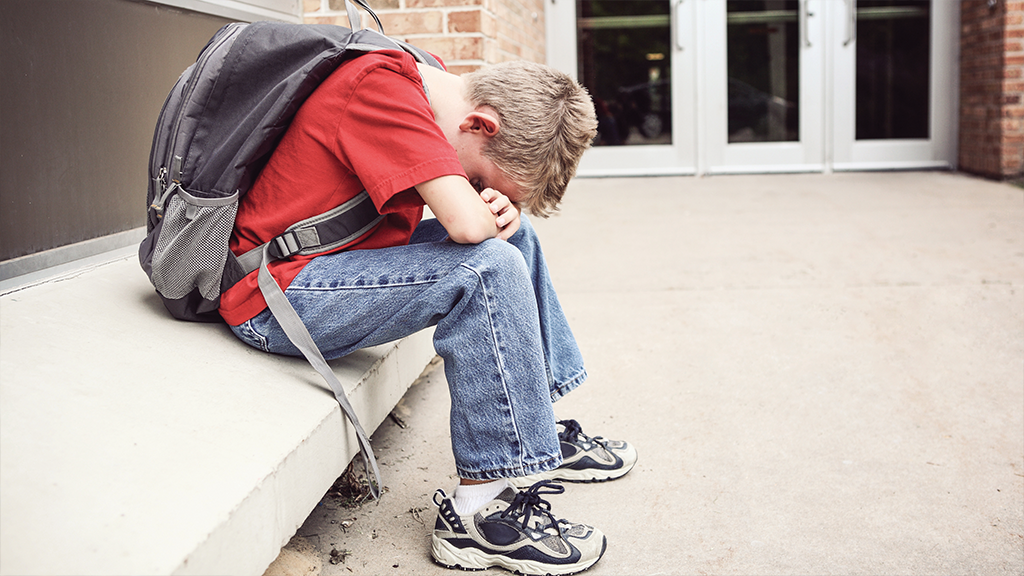 A young boy wearing a backpack puts his head on his hands and knees and looks sad. 