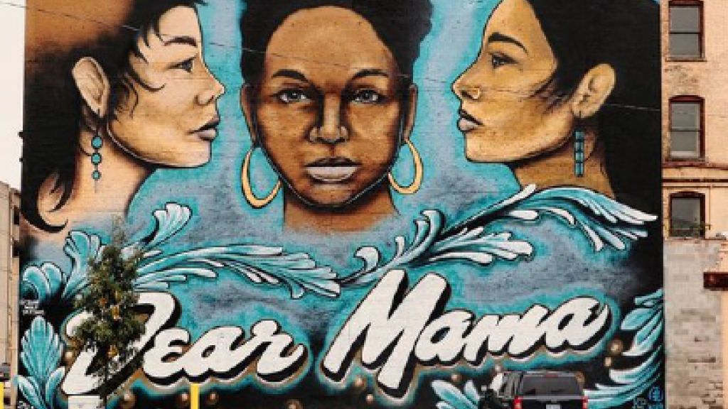 A mural painted on the side of building shows three women of different races and ethnicities and has the words Dear Mama written on the bottom