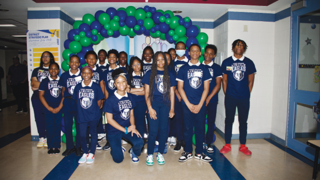 15 to 20 middle school students dressed in matching blue shirts stand under an arch made of balloons.