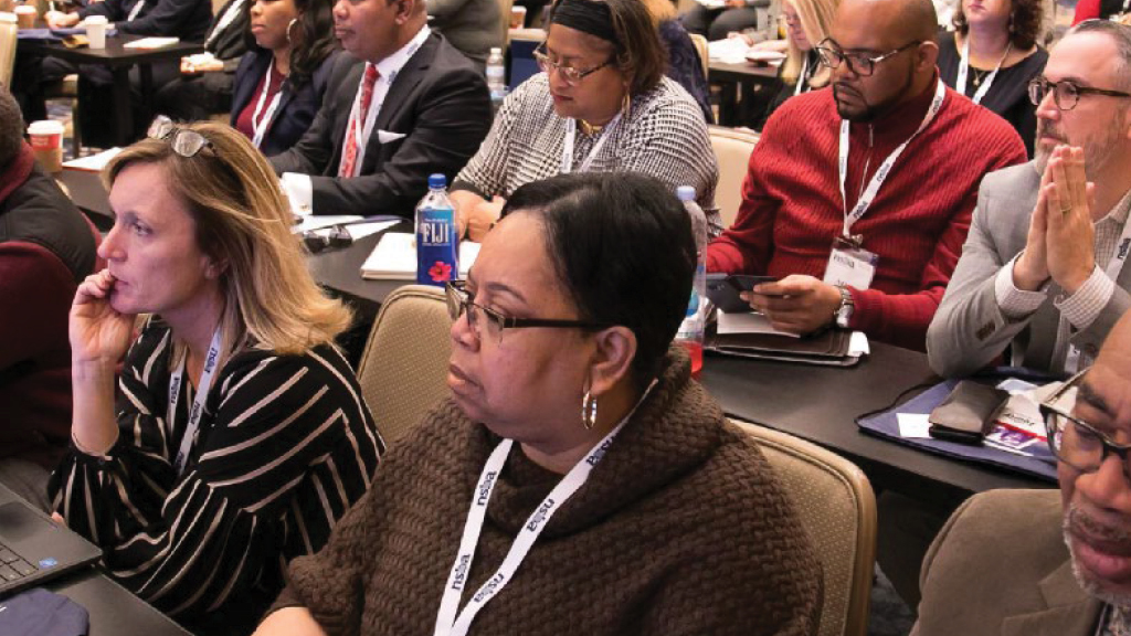 a diverse group of adults sit at a conference taking notes 